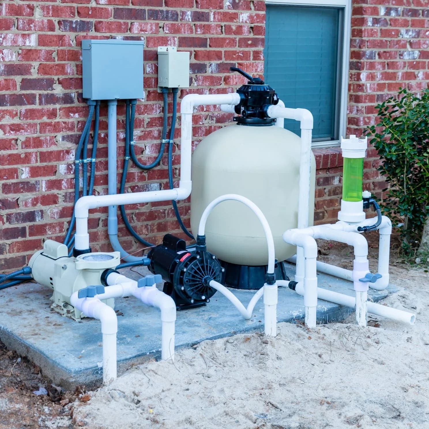 Residential pool pump and filter system with pipes, mounted on a concrete slab against a brick wall.